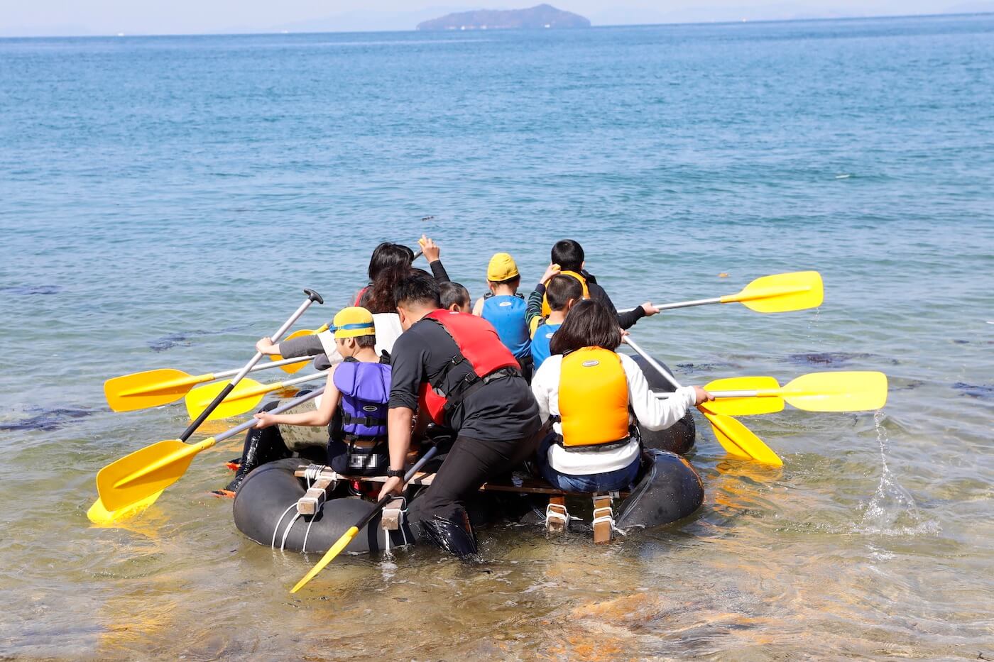 写真:一隻のイカダに乗って海へ漕ぎ出す複数の子どもと大人