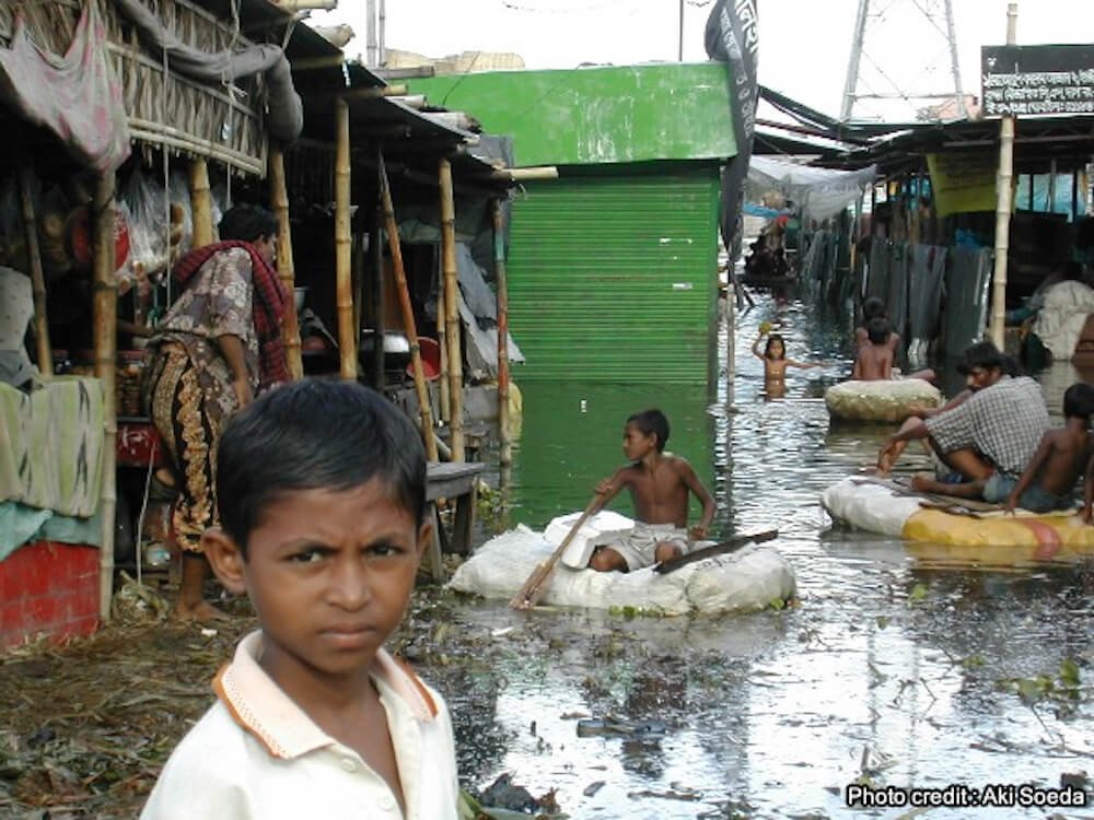 写真:洪水で浸水した村に立つ子ども。その後ろに複数の大人や子どもが浸水した村の中で暮らす様子