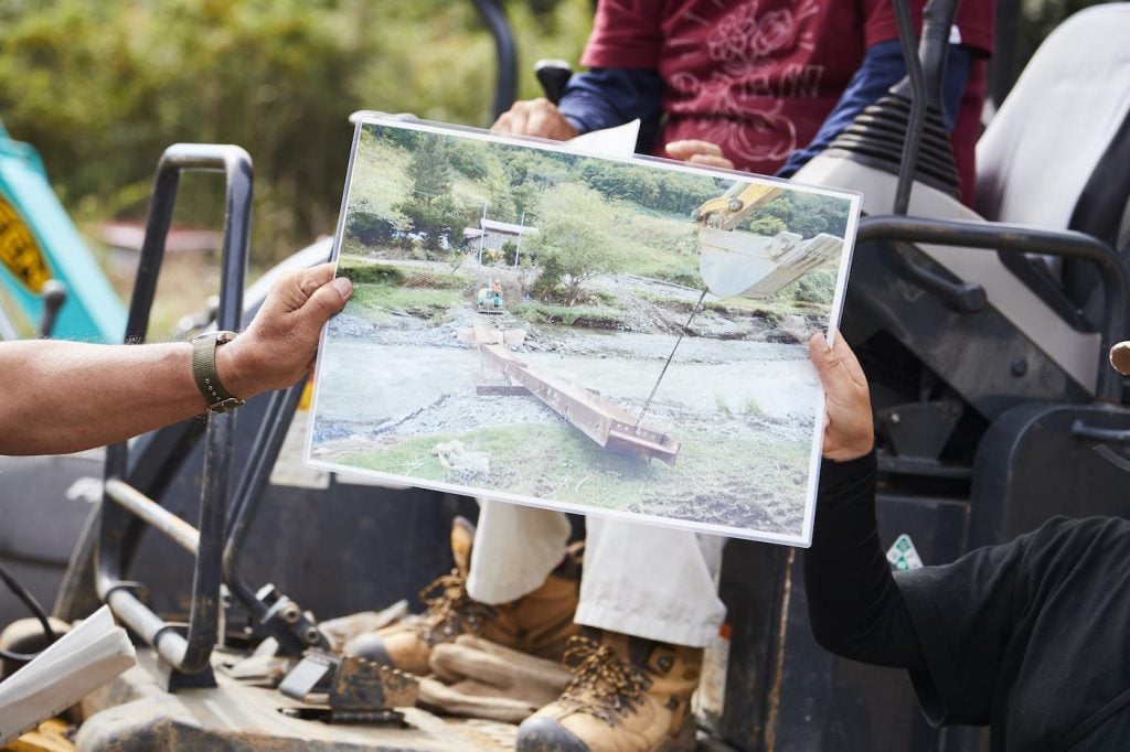 写真:川に倒壊した橋の代わりに簡易版の鉄の橋をかける小型重機