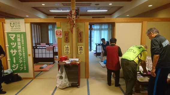 写真:温泉施設内にある畳の部屋の入り口付近で、マッサージの順番を待つ女性やスタッフの後ろ姿