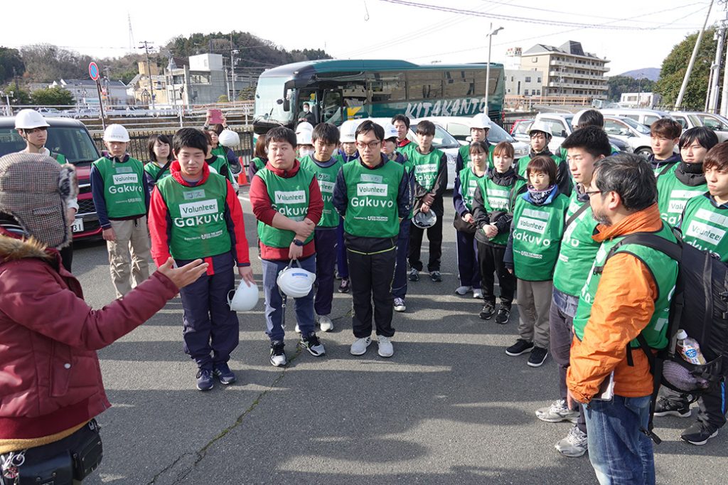 写真:駐車場で担当者からボランティアの注意事項などを説明される大学生たち