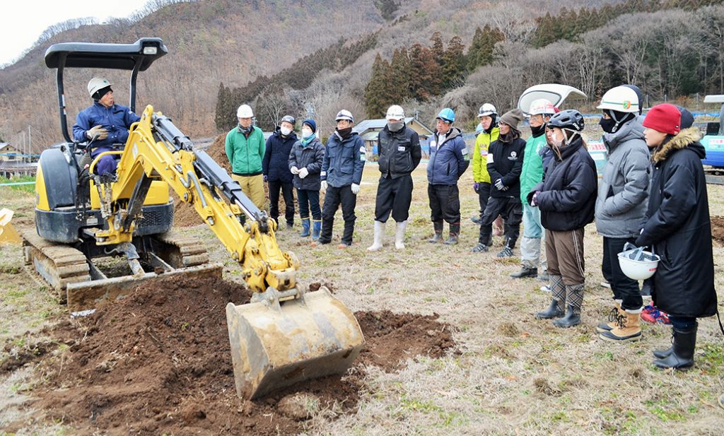 写真:ショベルを囲むように並び、参加者たちが講師の説明を聞く様子