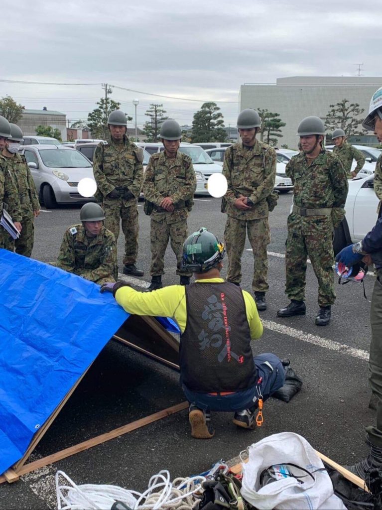 写真:屋根の模型を使ってブルーシートの張り方を説明する愛・知・人のメンバーと、その説明を聞く自衛隊員の様子