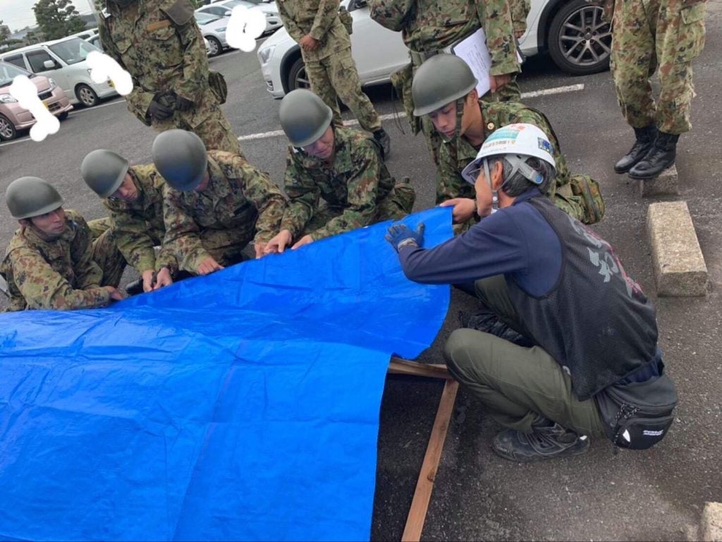 写真:屋根の模型を使ってブルーシートの張り方を説明する愛・知・人のメンバーと、その説明を聞く自衛隊員の様子