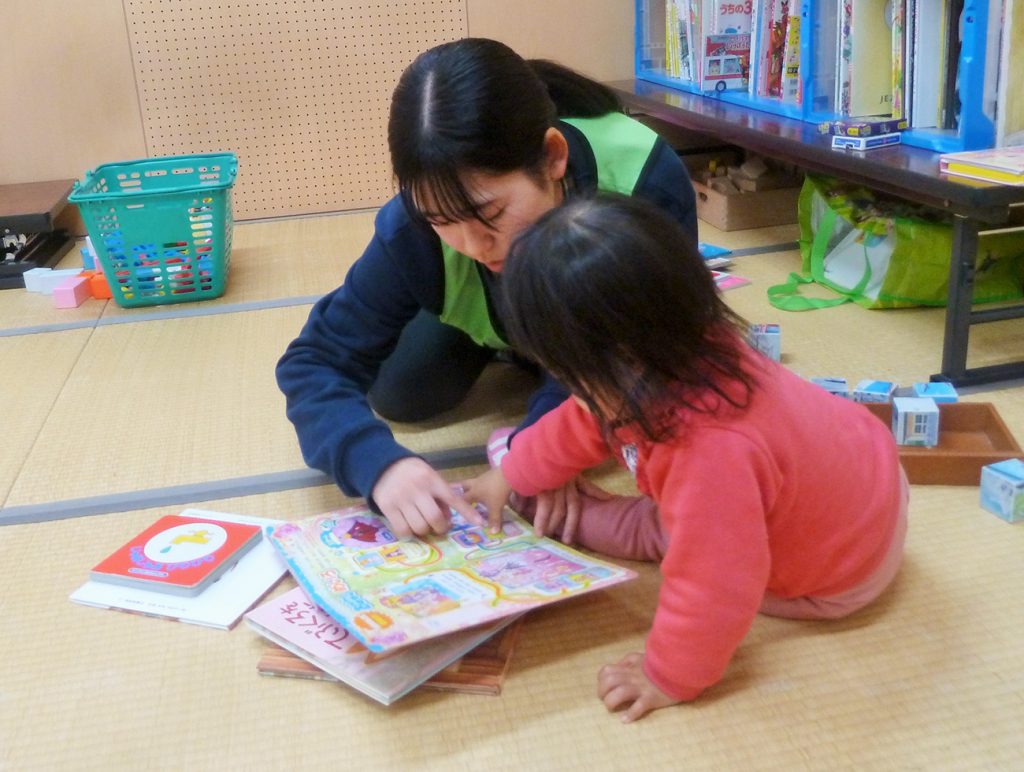 写真:畳の上に座り、被災地の子どもに指で追いながら絵本の読み聞かせをする高校生の様子(12月8日、横沢祥子さん提供)