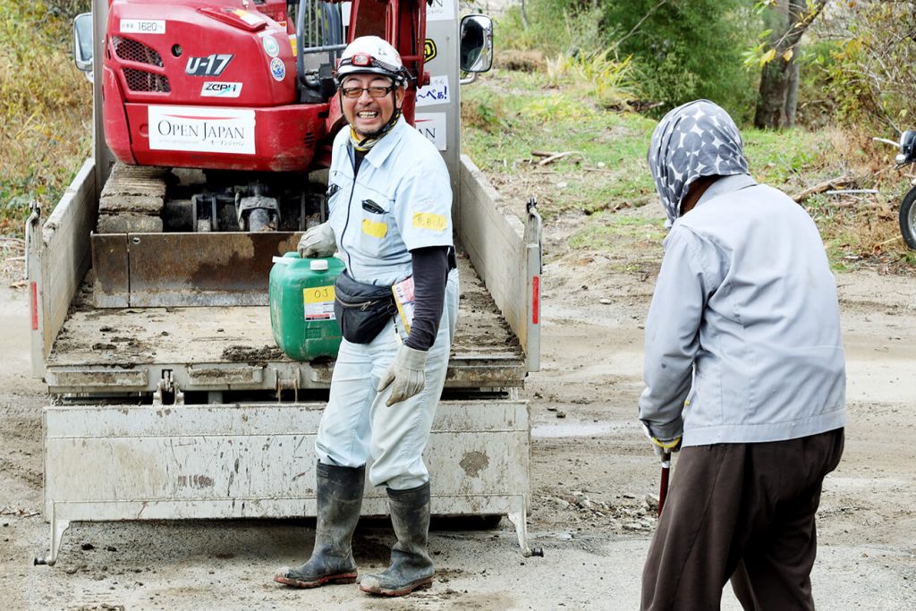 写真：丸森町で重機を背に、作業服姿で住民と話す萬代さん（左）（OPEN JAPAN提供）