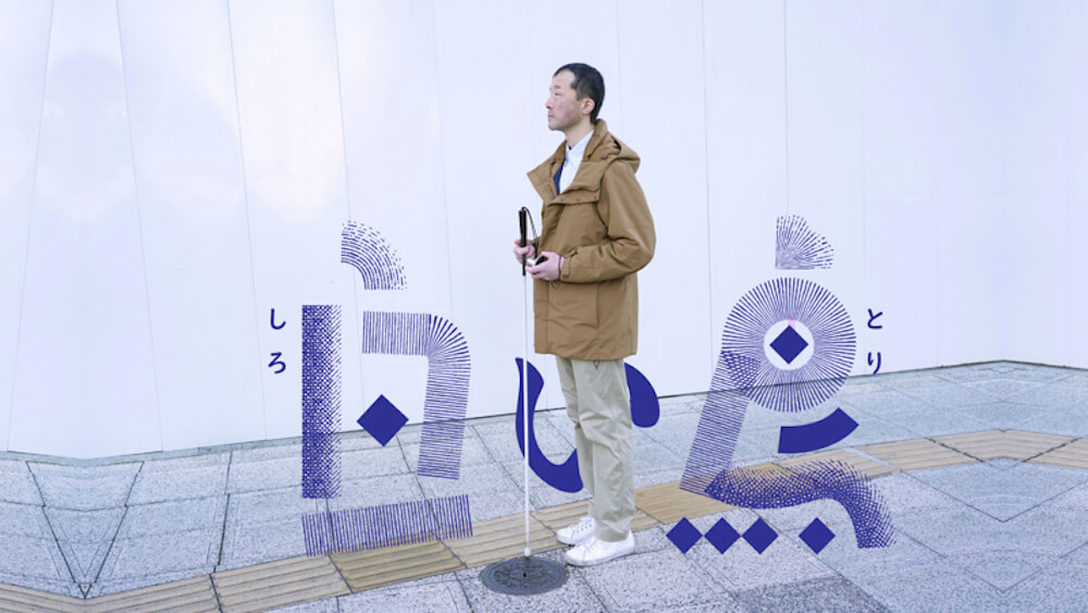 画像:ドキュメンタリー映画「白い鳥」のワンシーン。歩道の上に立つ白状を持った視覚障害者