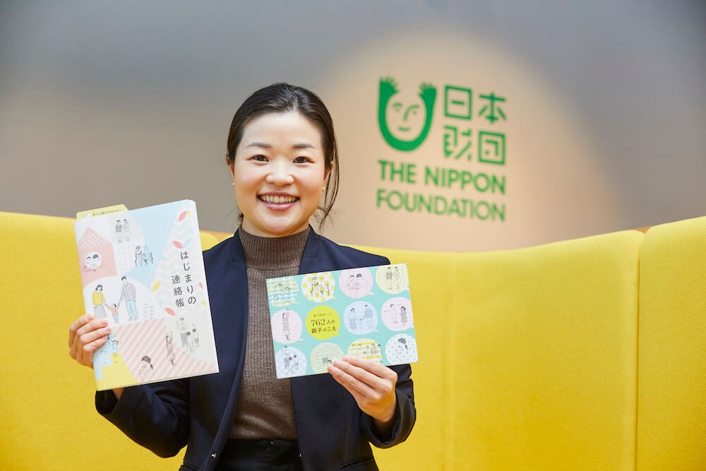写真:日本財団が作成した冊子「はじまりの連絡帳」「養子縁組をした762人の声」を手に持ち微笑む新田さん