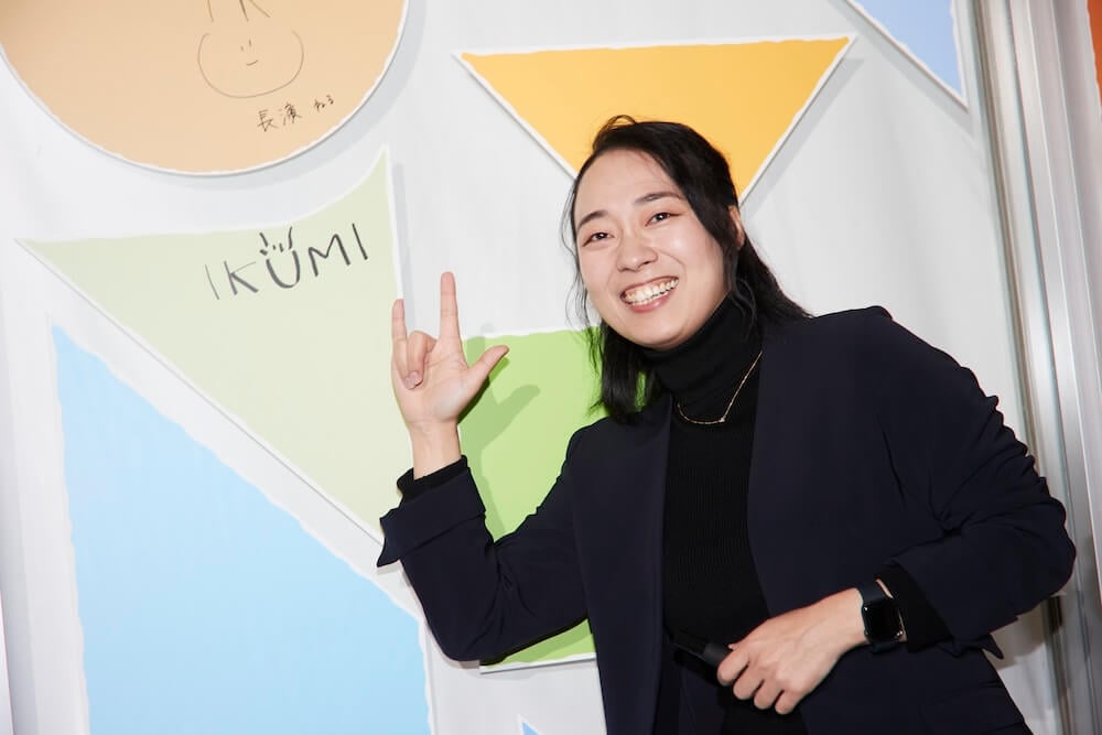 画像：「みるカフェ」の壁に書いた自らのサインの前で、「アイ・ラブ・ユー」と手話でポーズをよる川俣さん