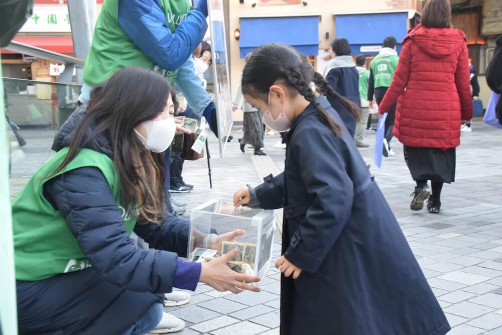 写真:ボランティアが持つ募金箱にお金を寄付する少女