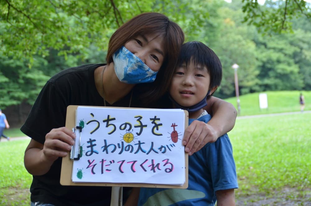 公園の中で「うちの子をまわりの大人がそだててくれる」というボードを持った母子