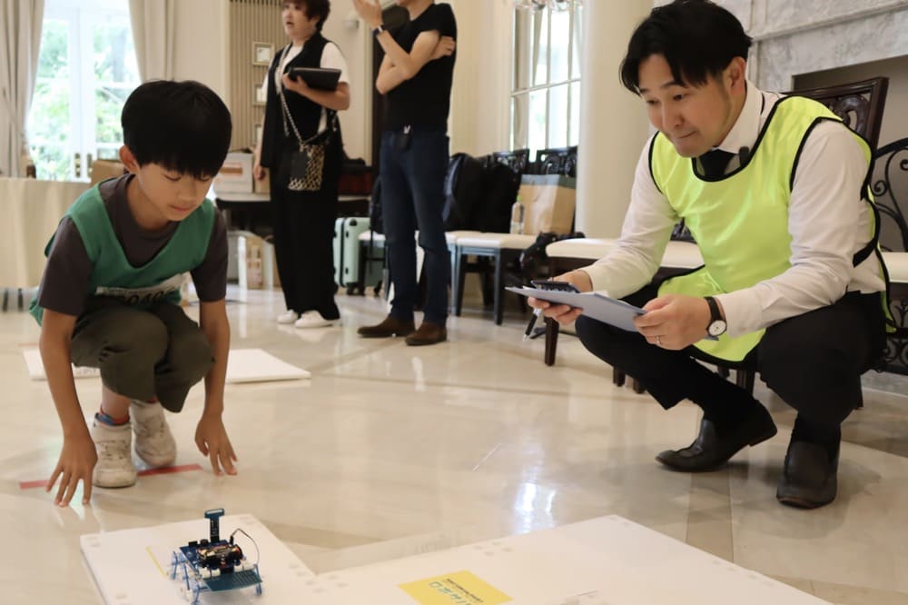 自分が作ったロボットを操作する子どもと、それを審査するスタッフ
