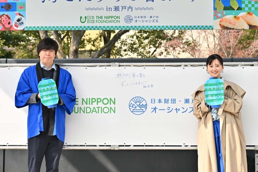 写真：メッセージフラッグの前で自筆のメッセージの前に立つ飯沼さんと日本財団スタッフ