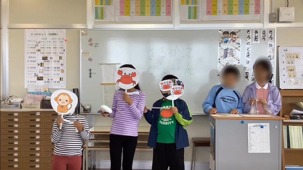 「さるかにがっせん」の寸劇に取り組む子どもたち。
3人の生徒が、猿や蟹のイラストが貼られたうちわを顔の前にかざし、それぞれの役を演じている。教壇のそばでは、2人の生徒がその様子を見守っている。