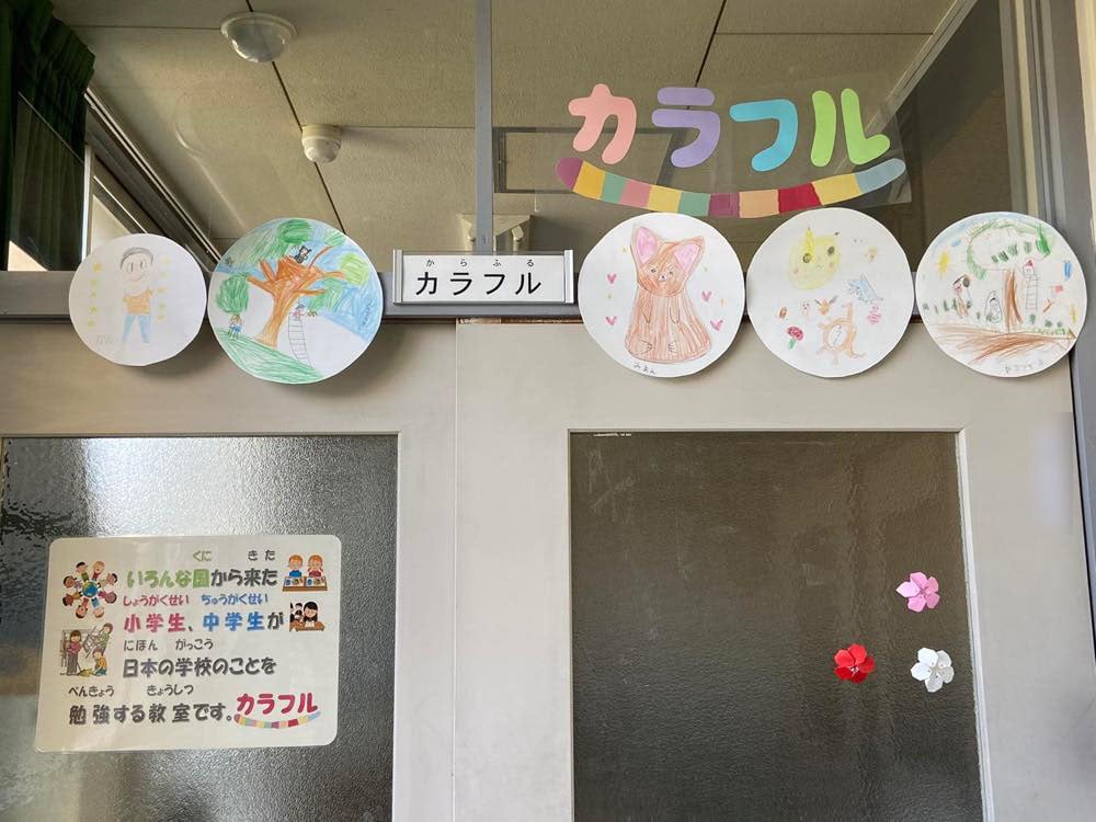 カラフルの教室の入り口。
ガラス格子の引き戸には「いろんな国から来た小学生、中学生が日本の学校のことを勉強する教室です。」という案内が貼られている。まわりには、生徒が描いたと思われるイラストや、「カラフル」の文字を切り抜いた画用紙、折り紙の花などが飾られている。