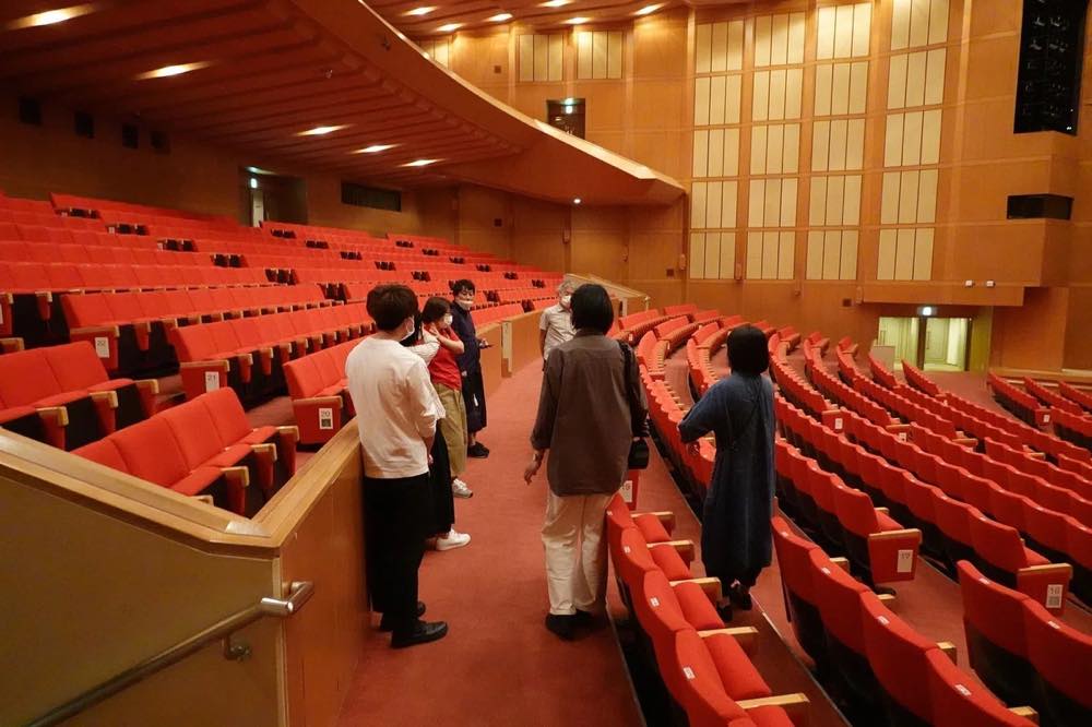 福岡県大牟田市にある大牟田文化会館を、プロジェクトのメンバーが見学している。舞台に向かって赤い客席が並ぶ、公演会場のような空間。