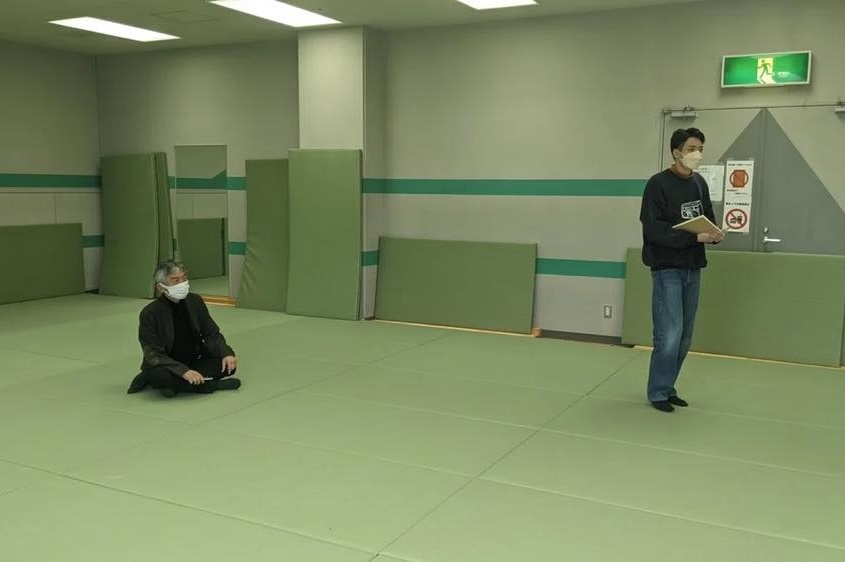 宮城県岩沼市の総合体育館体育館。緑色のマットが敷き詰められた床に櫛名誉教授が座っている。