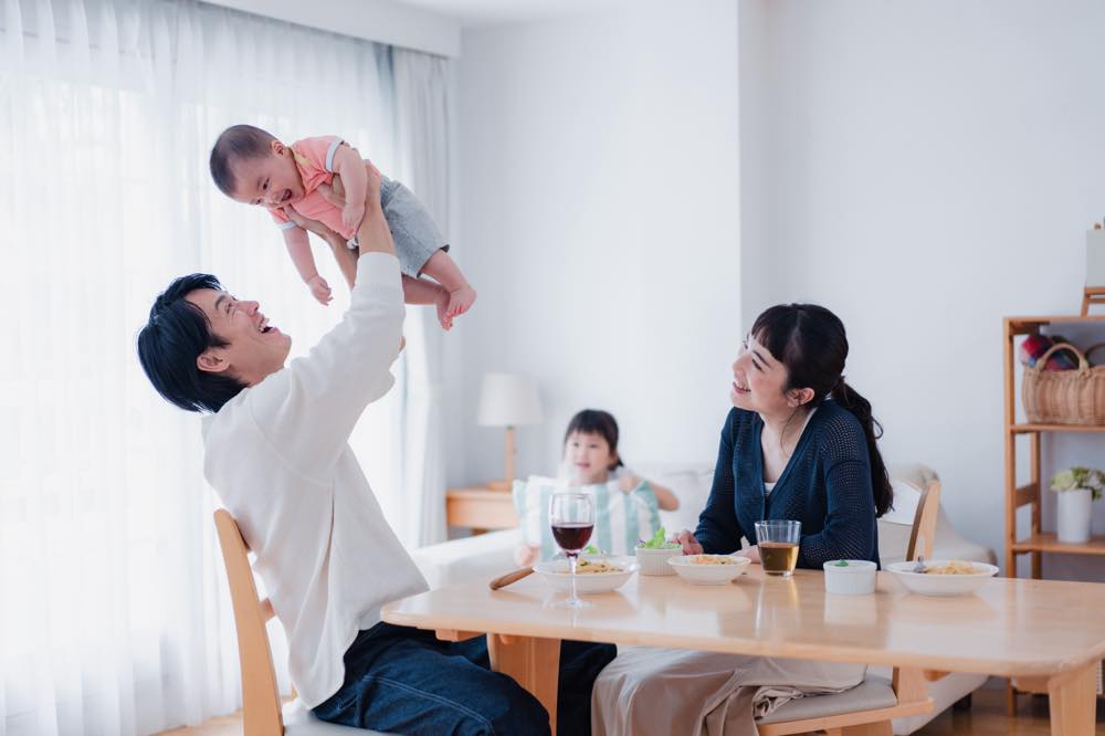 リビングに、夫婦(男女)と乳児、幼児がいる。父親と見られる男性が乳児を「たかいたかい」している