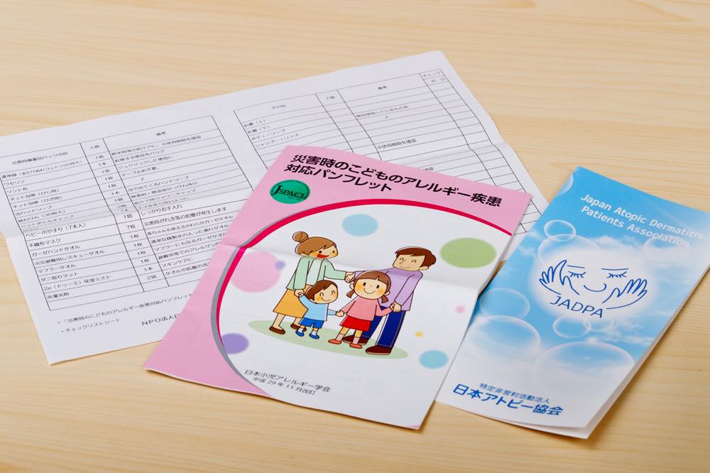 日本アトピー協会がアトピー患者向けに制作した「災害時備蓄品リスト」と、日本アトピー協会のパンフレットと、日本小児アレルギー学会の「災害時の子どものアレルギー疾患対応パンフレット」の写真