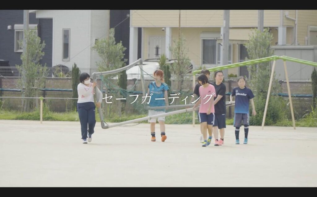 S.C.P.JapanのYouTubeで公開されている動画「スポーツにおけるセーフガーディング（コーチの声編）」のキャプチャー。屋外のグラウンドで、子どもとスタッフがサッカーゴールのフレームを運んでいる様子。画面中央には大きく「セーフガーディング」という白い文字が書かれている。