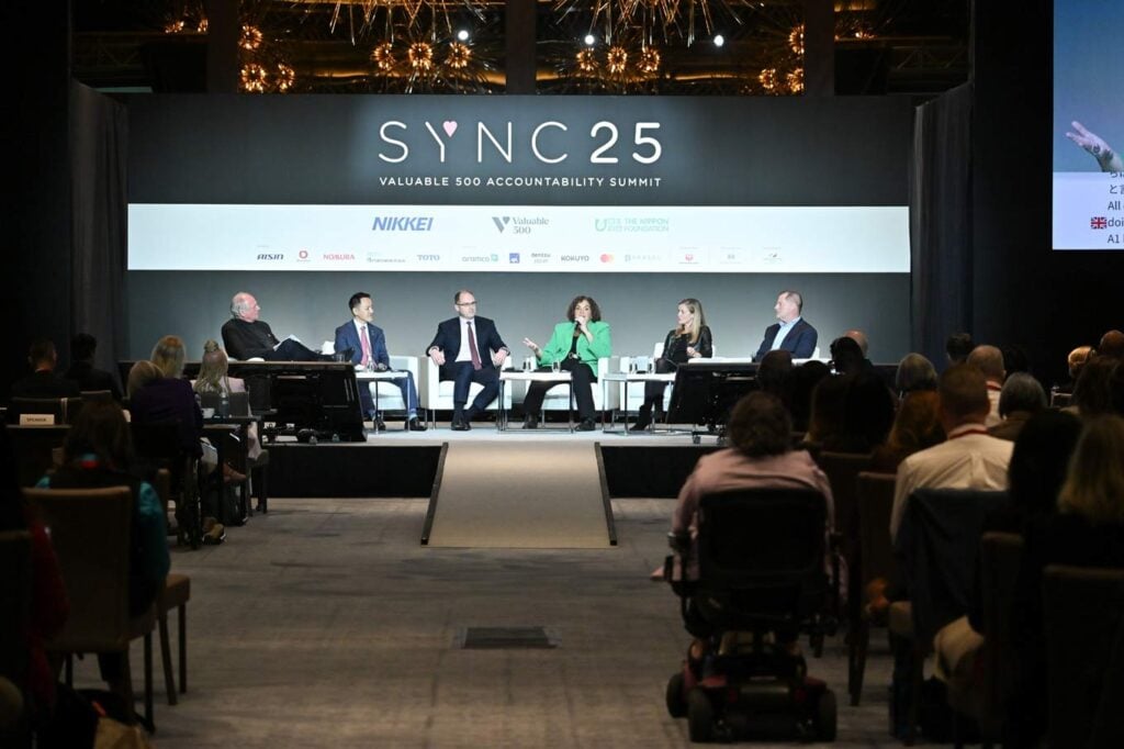 写真:SYNC25アカウンタビリティ・サミットで、世界的大企業の経営幹部複数名がステージ上で障害者インクルージョンについてパネルディスカッションを行っている様子。客席には車椅子利用者を含む参加者が座っている。)