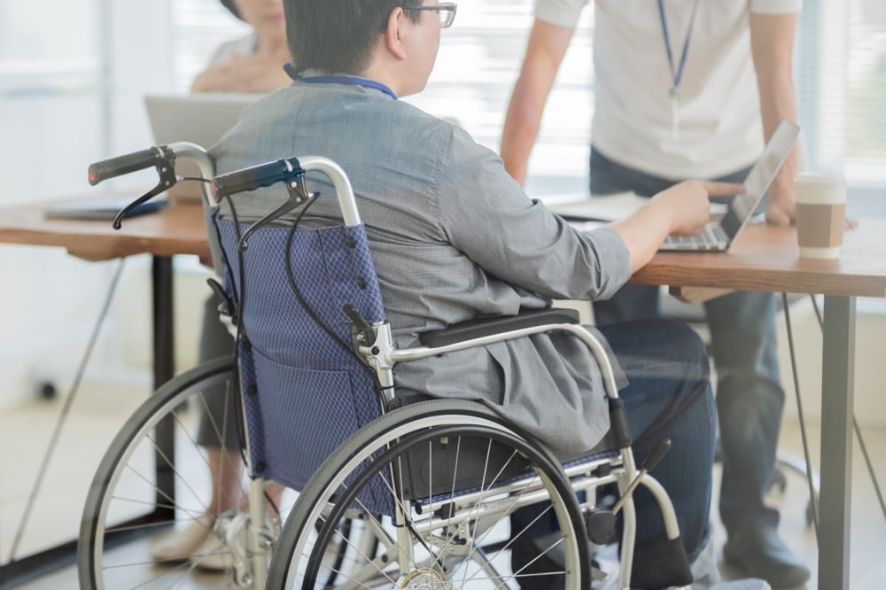 オフィスで車椅子に座りながら、同僚と一緒にパソコンの画面を覗き込んで仕事をしている男性の背後からの写真。